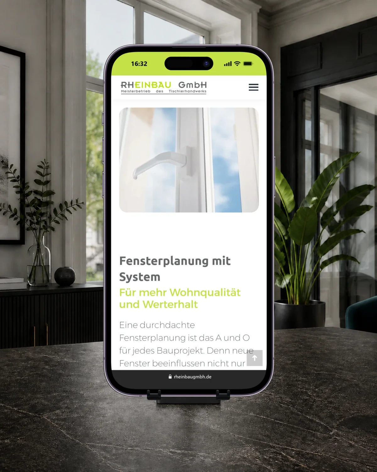 iPhone auf einer Halterung mit der Rheinbau-Seite zu Kunststofffenstern und dem Abschnitt „Fensterplanung mit System“