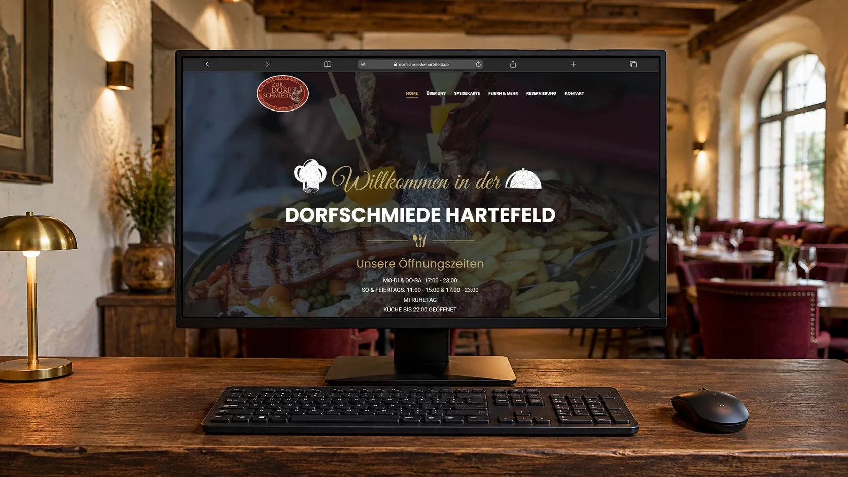 Desktop-Monitor mit der Startseite der Dorfschmiede Hartefeld, Tastatur und Maus auf einem Holztresen in einer Restaurant-Szene.