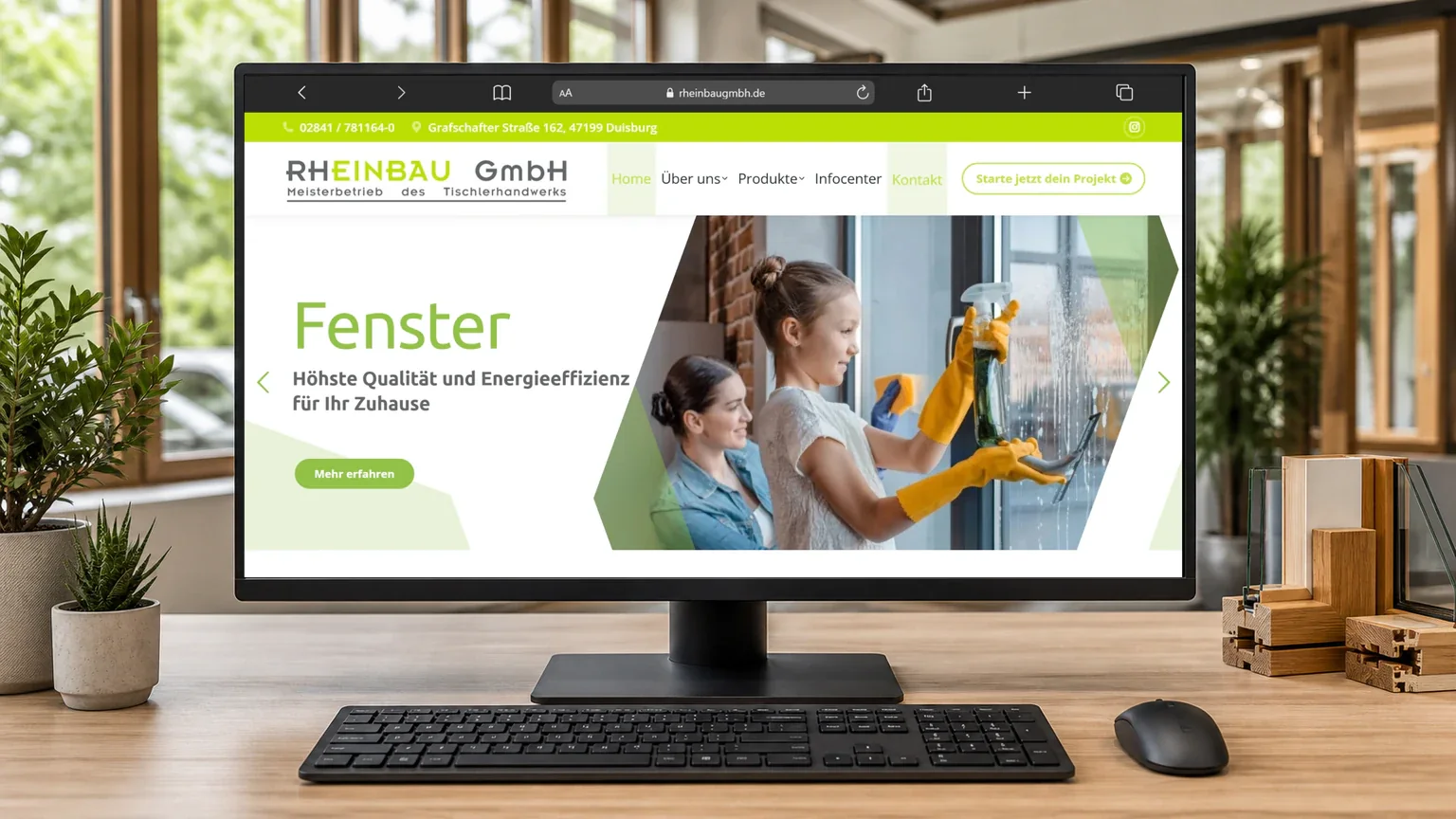 Monitor mit der Rheinbau-Website zum Thema Fenster, davor Tastatur und Maus in einer hellen Arbeitsumgebung mit Fensterprofilen