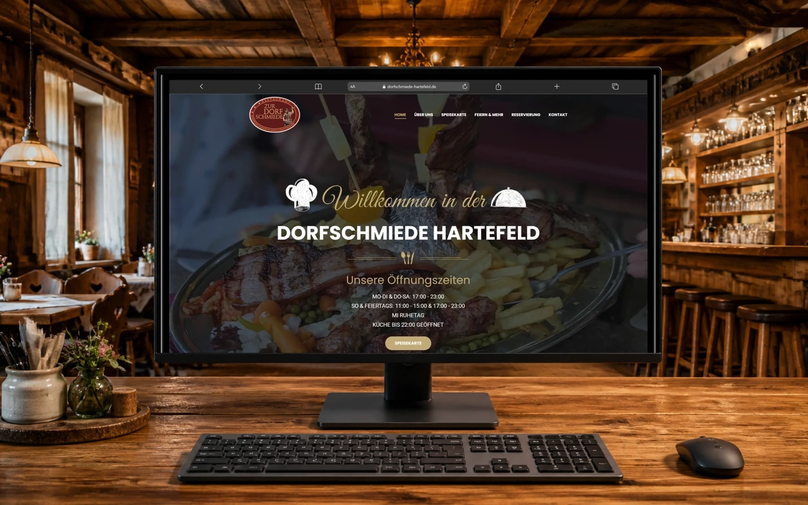 Desktop-Monitor mit der Startseite der Dorfschmiede Hartefeld; davor liegen Tastatur und Maus auf einem Holztisch.